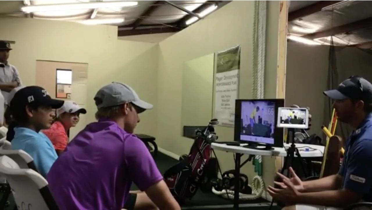 Junior golfers seated in an indoor bay watching swing video on a monitor while their coach talks them through it